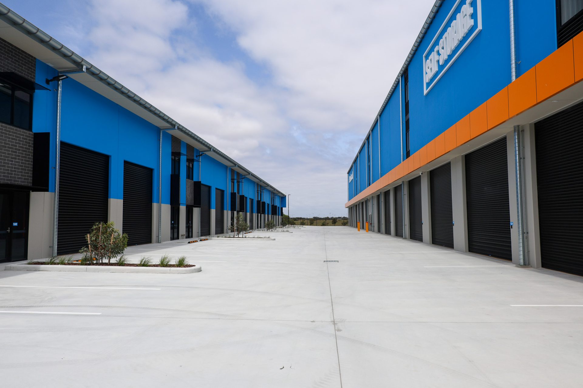 A wide driveway between two rows of modern industrial storage buildings, featuring blue and gray facades with large black roller doors. A sign reading 'Self Storage' is displayed on the right building, and sparse landscaping is visible along the driveway.