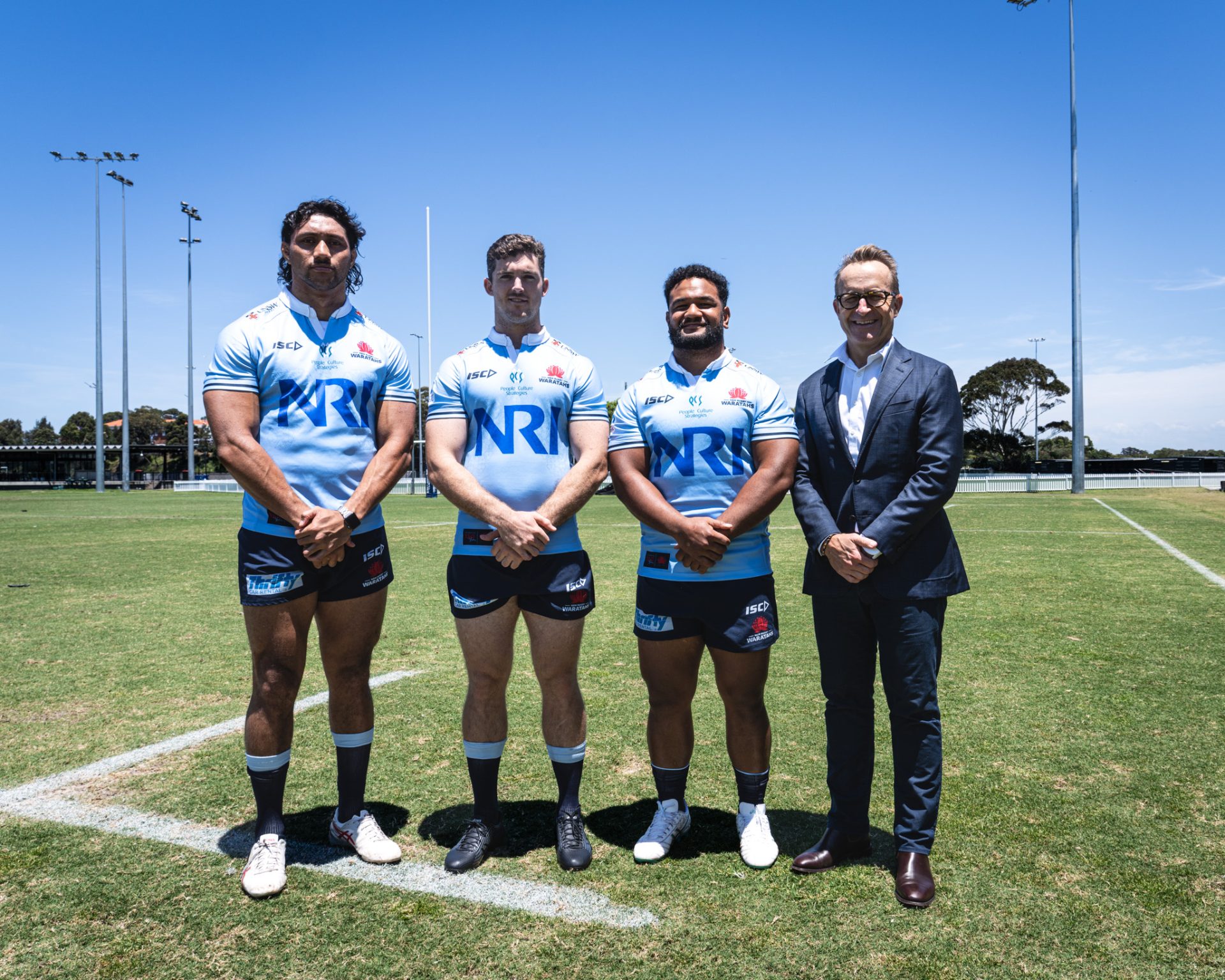 Four individuals standing on a rugby field, including three NSW Waratahs rugby players wearing light blue jerseys with 'NRI' sponsor logos, black shorts, and socks. The players stand with their hands clasped in front, alongside a man in a dark suit on the right, likely a team official or sponsor representative. The scene is set under a clear blue sky, with rugby goalposts, green grass, and a few trees visible in the background